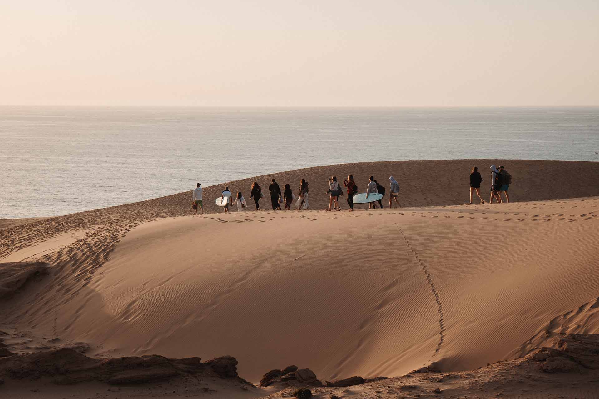 Un gruppo SurfWeek cammina sul crinale di una duna verso l'oceano al tramonto, con le tavole sotto il braccio