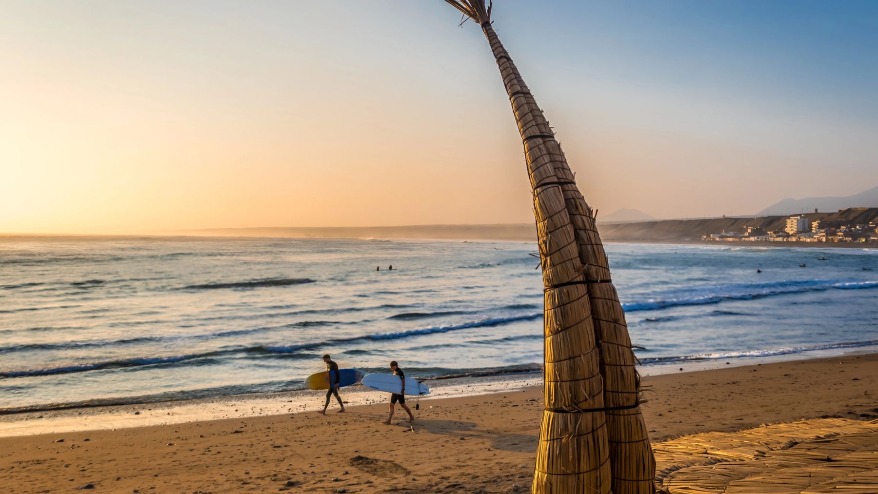 Due surfisti camminano sulla spiaggia di Huanchaco accanto a un caballito de totora