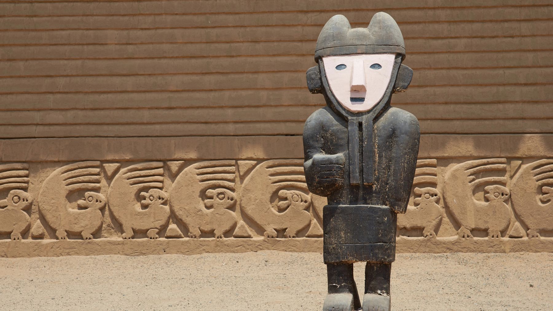 Una statua in legno davanti ai fregi di fango di Chan Chan